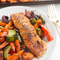 White plate with a salmon filet on it and roasted veggies next to it. Sheet pan of veggies in the background.