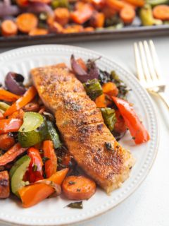 White plate with a salmon filet on it and roasted veggies next to it. Sheet pan of veggies in the background.