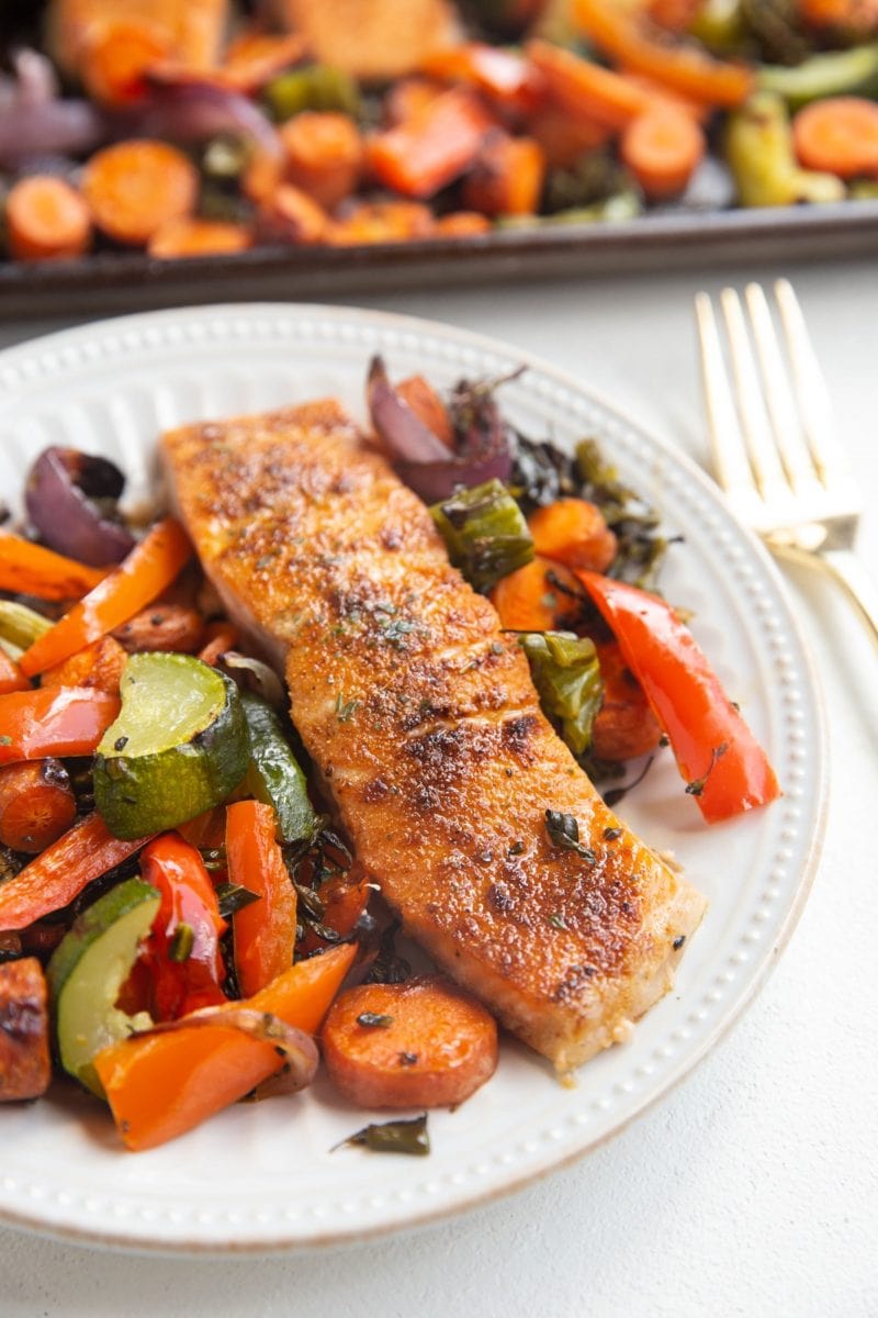 White plate with a salmon filet on it and roasted veggies next to it. Sheet pan of veggies in the background.