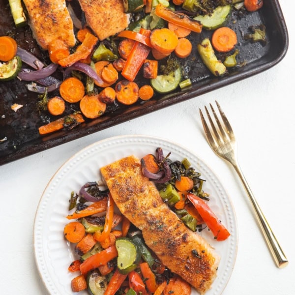 top-down photo of a sheet pan with salmon and vegetables and a white plate with salmon and vegetables with a gold fork next to it.