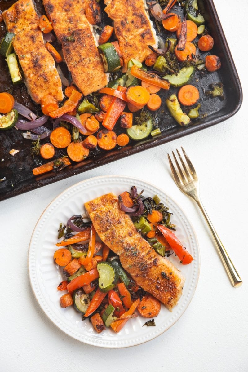 top-down photo of a sheet pan with salmon and vegetables and a white plate with salmon and vegetables with a gold fork next to it.
