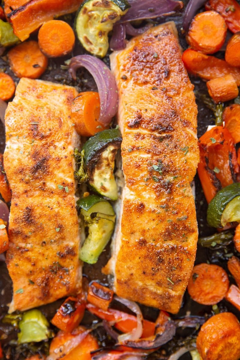 Close up shot of juicy cooked salmon on a sheet pan with vegetables surrounding it.