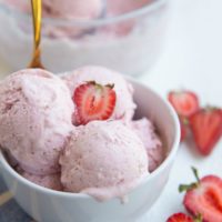 White bowl of strawberry ice cream with a gold spoon and fresh strawberries to the side.