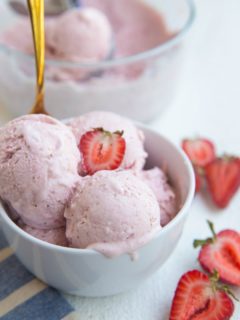 White bowl of strawberry ice cream with a gold spoon and fresh strawberries to the side.