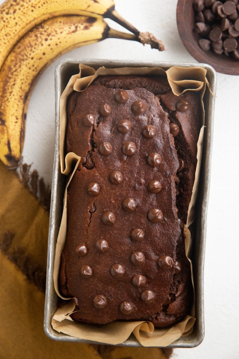 Loaf of chocolate banana bread in a baking pan with two ripe bananas to the side, a bowl of chocolate chips, and a golden napkin.