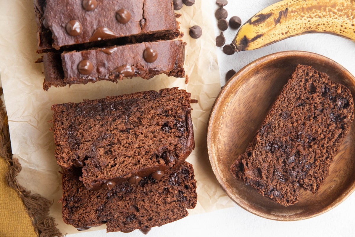 Horizontal image of sliced loaf of banana bread with a wooden plate to the side with a slice on top.