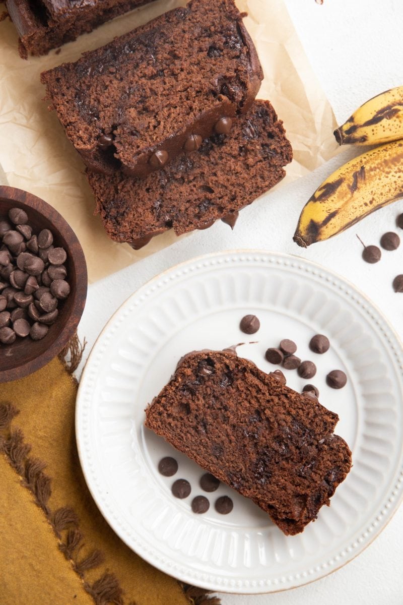 Loaf and slices of banana bread with a white plate with a slice of chocolate banana bread on top and bananas to the side.