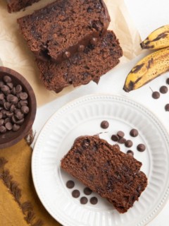 Loaf and slices of banana bread with a white plate with a slice of chocolate banana bread on top and bananas to the side.