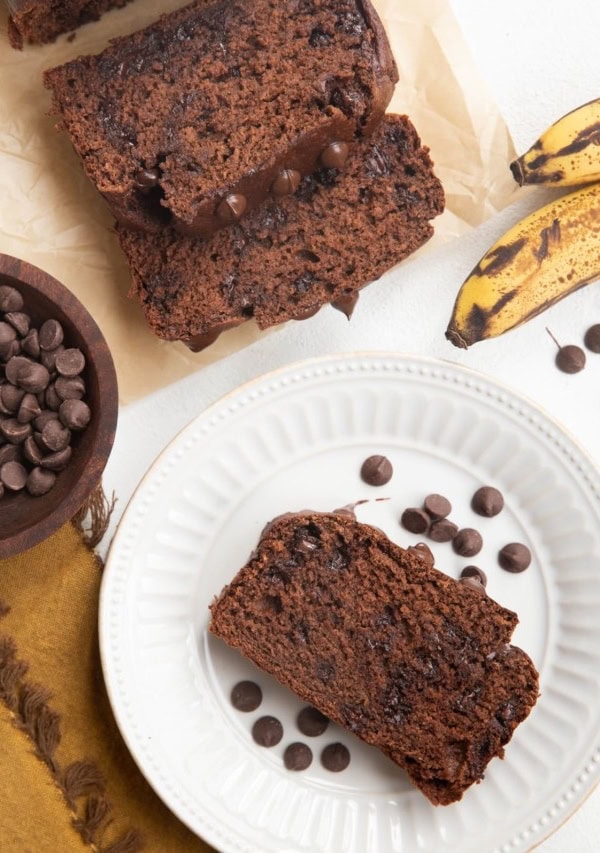 Loaf and slices of banana bread with a white plate with a slice of chocolate banana bread on top and bananas to the side.