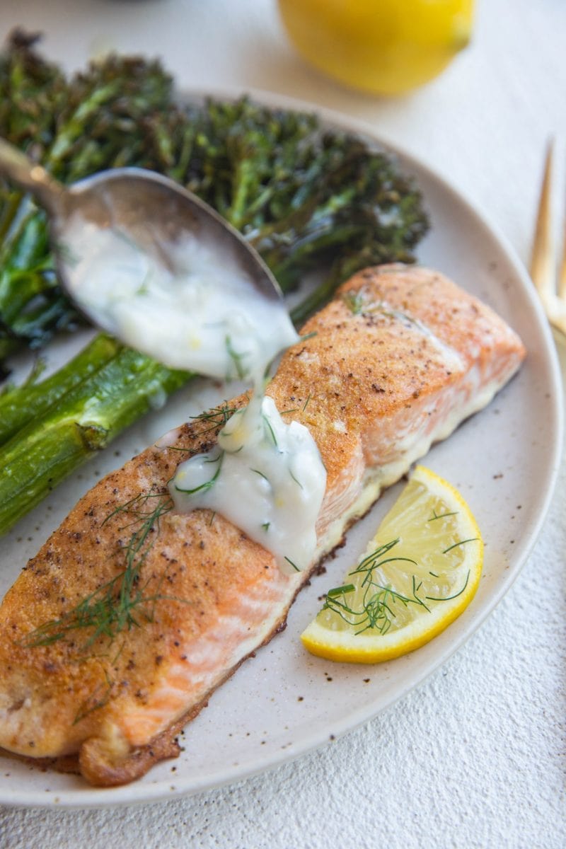 baked salmon on a plate with broccolini and dill sauce being poured on top.