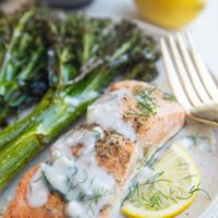 Baked salmon on a plate with dill sauce, broccolini and a lemon slice.