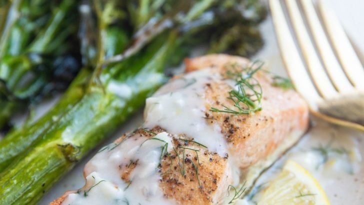 Baked salmon on a plate with dill sauce, broccolini and a lemon slice.