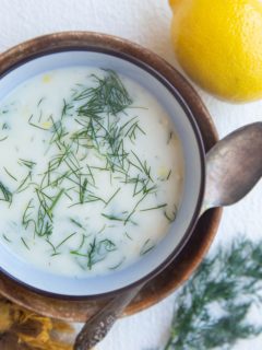 Creamy dill sauce in a blue bowl on top of a wooden plate. A silver spoon, lemon, and fresh dill to the side.