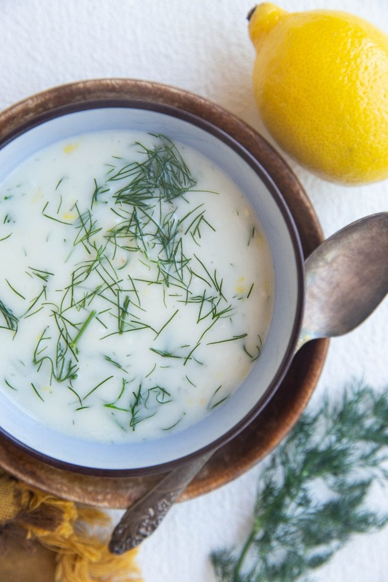 Creamy dill sauce in a blue bowl on top of a wooden plate. A silver spoon, lemon, and fresh dill to the side.