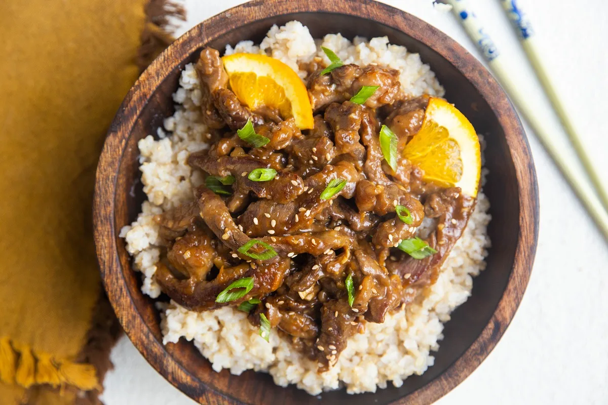 horizontal photo of orange Chinese beef in a wooden bowl with brown rice, sprinkled with chopped green onions.