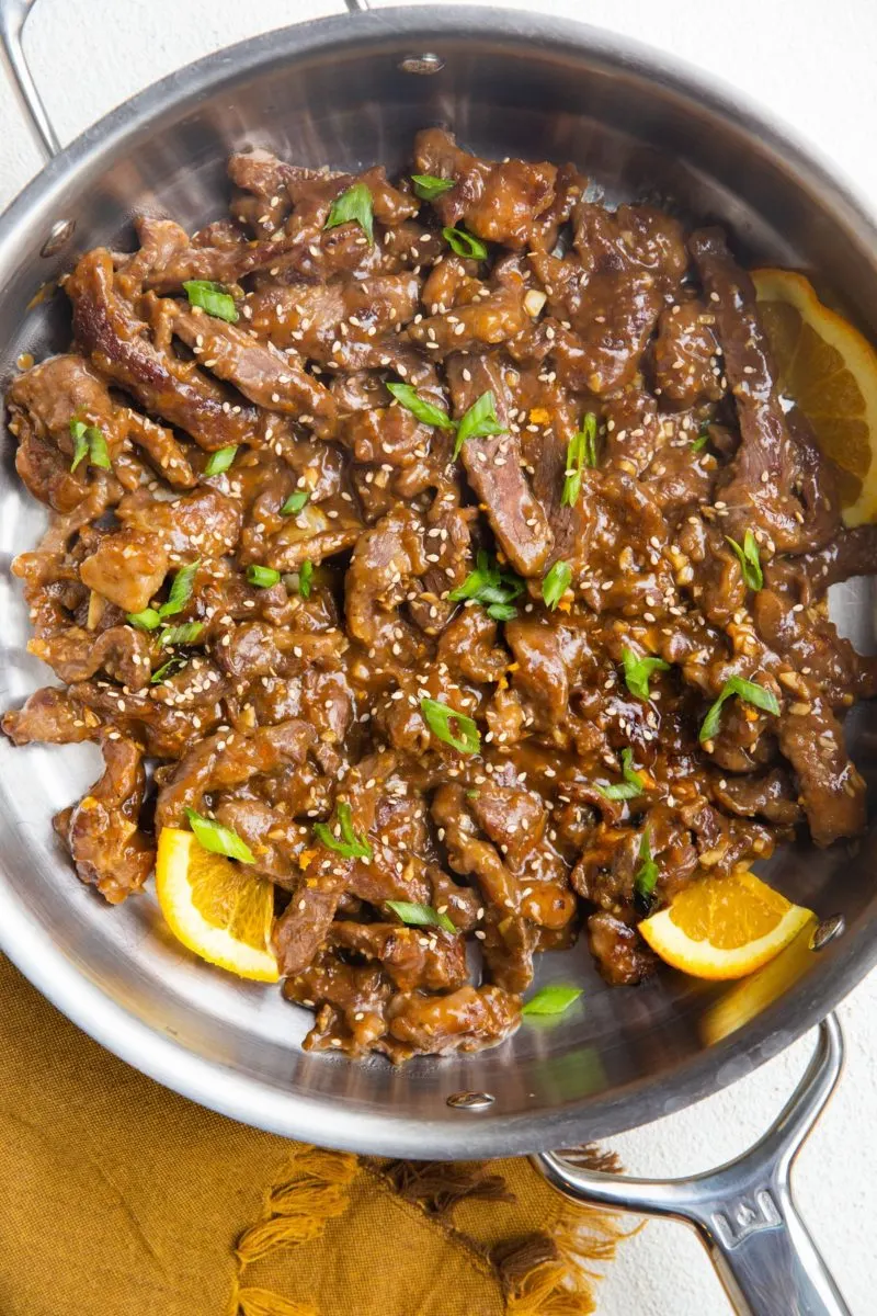 Stainless steel skillet with sesame ginger beef, chopped green onions and slices of orange.