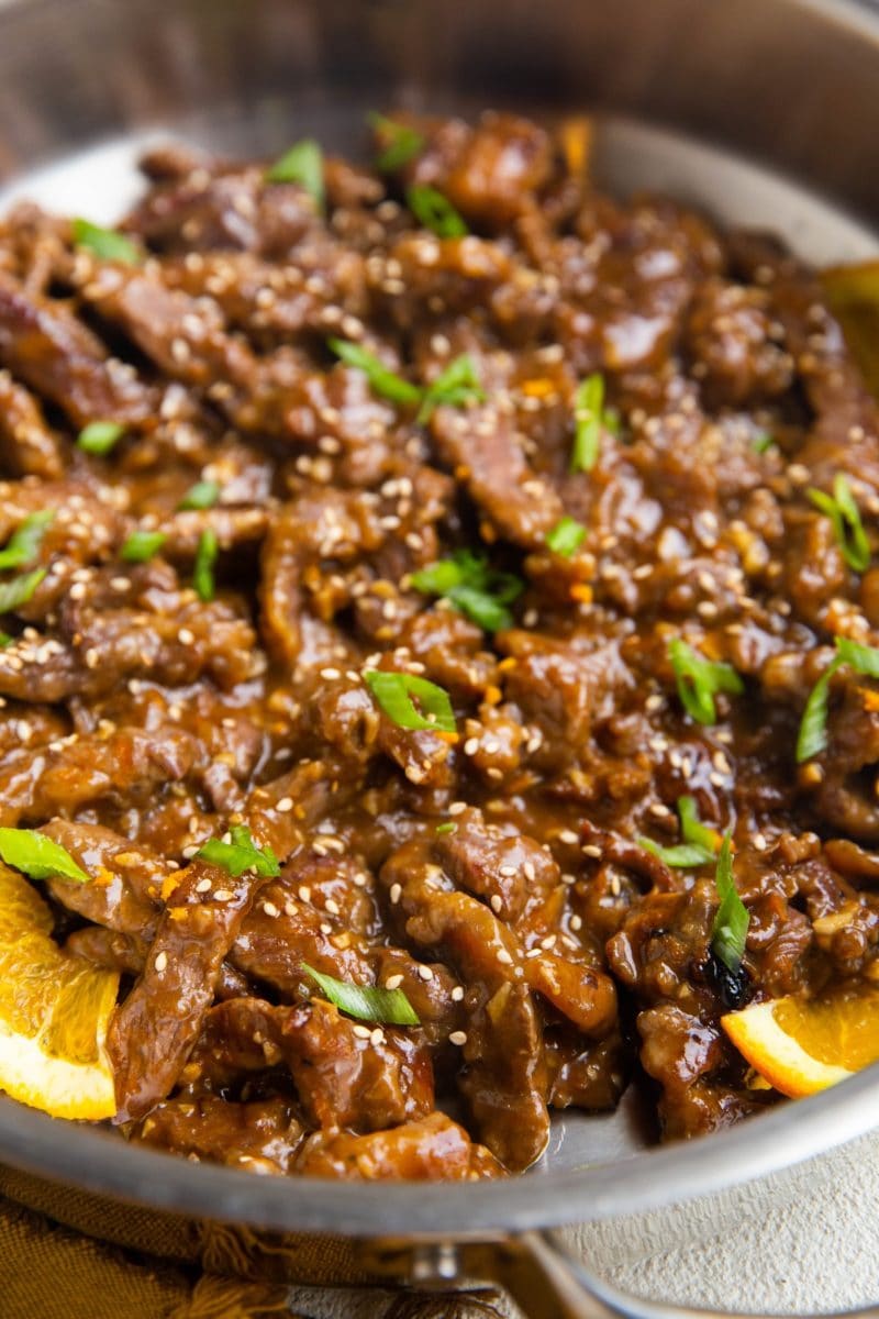 Orange beef in a stainless steel skillet with orange slices and green onions on top.