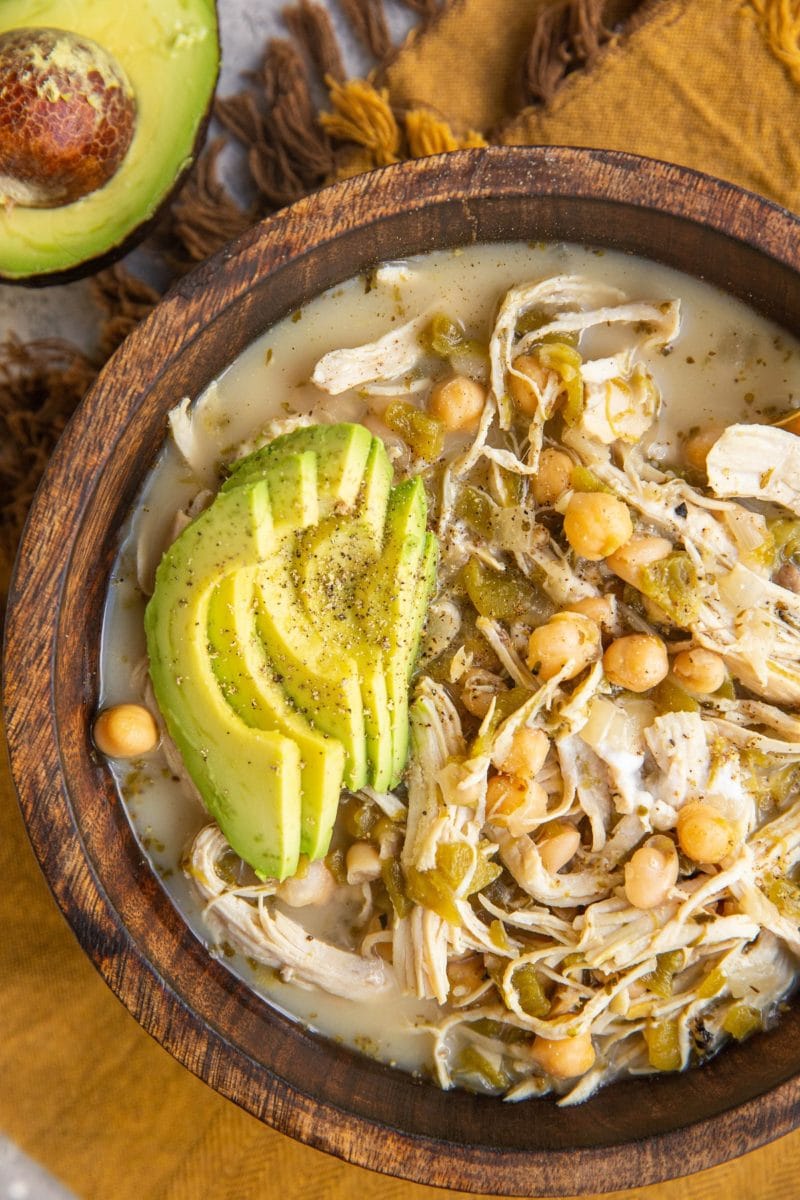 Wood bowl of chicken chili with sliced avocado on top. A golden napkin and half of an avocado to the side.