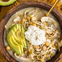 Big bowl of chili with Greek yogurt on top, sliced avocado and a gold spoon in the soup. A golden napkin underneath the bowl and half of an avocado to the side.