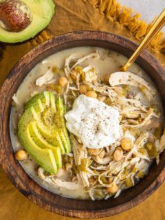 Big bowl of chili with Greek yogurt on top, sliced avocado and a gold spoon in the soup. A golden napkin underneath the bowl and half of an avocado to the side.