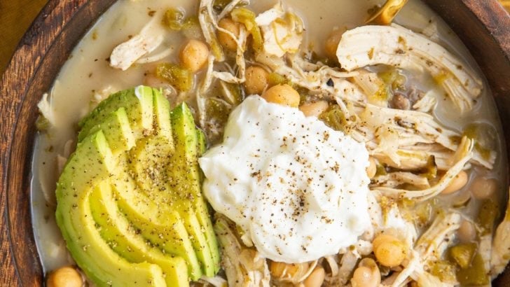 Big bowl of chili with Greek yogurt on top, sliced avocado and a gold spoon in the soup. A golden napkin underneath the bowl and half of an avocado to the side.