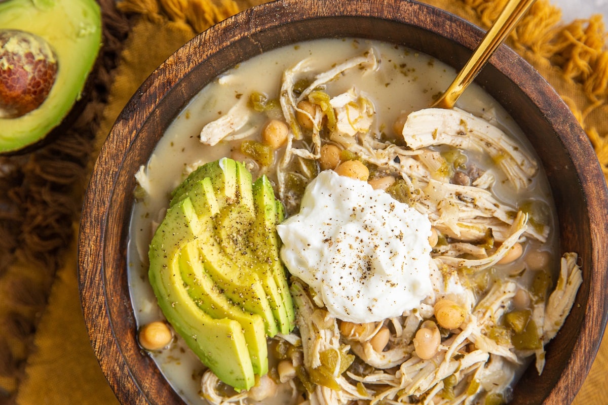 horizontal photo of white chicken chili in a wooden bowl with sour cream and avocado on top.