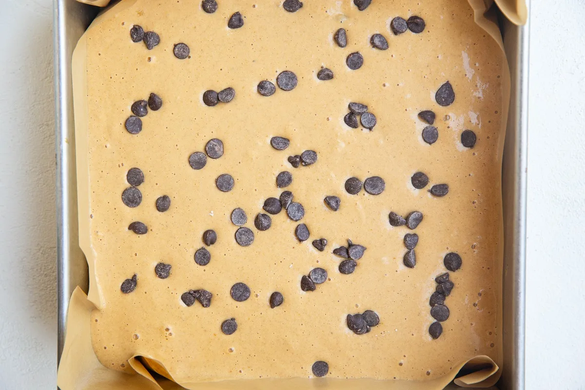 Batter in a parchment-lined square baking pan with chocolate chips on top.