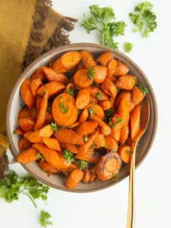 Roasted carrots in a bowl with a golden spoon, ready to serve. Fresh parsley to the side along with a golden napkin.