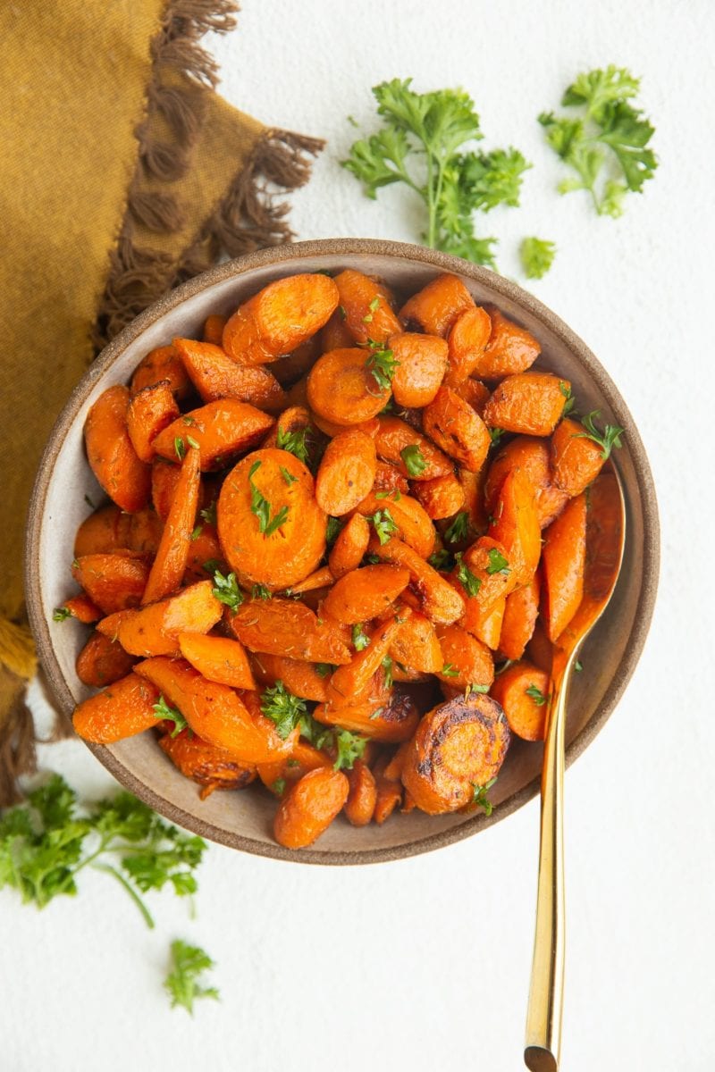 Roasted carrots in a bowl with a golden spoon, ready to serve. Fresh parsley to the side along with a golden napkin.