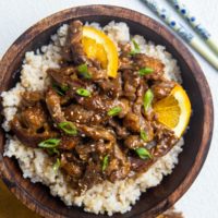 Big wooden bowl of brown rice and sesame ginger orange beef with chopsticks to the side.