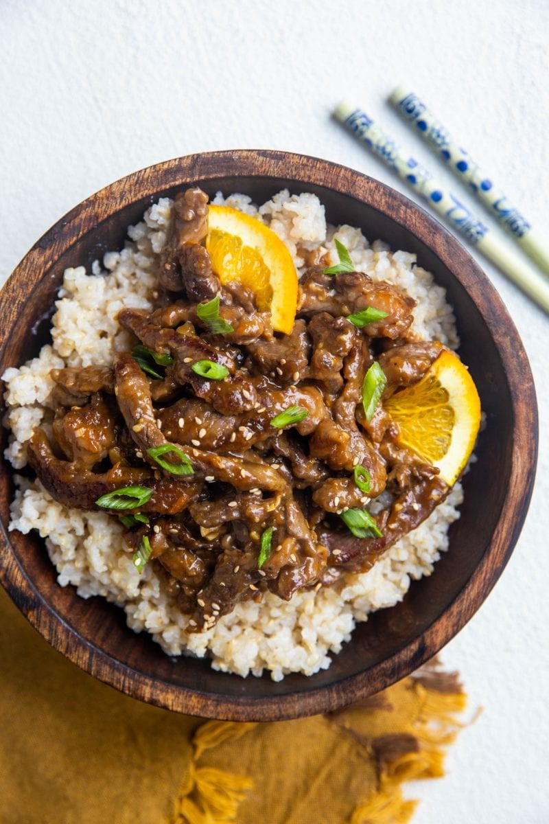 Big wooden bowl of brown rice and sesame ginger orange beef with chopsticks to the side.