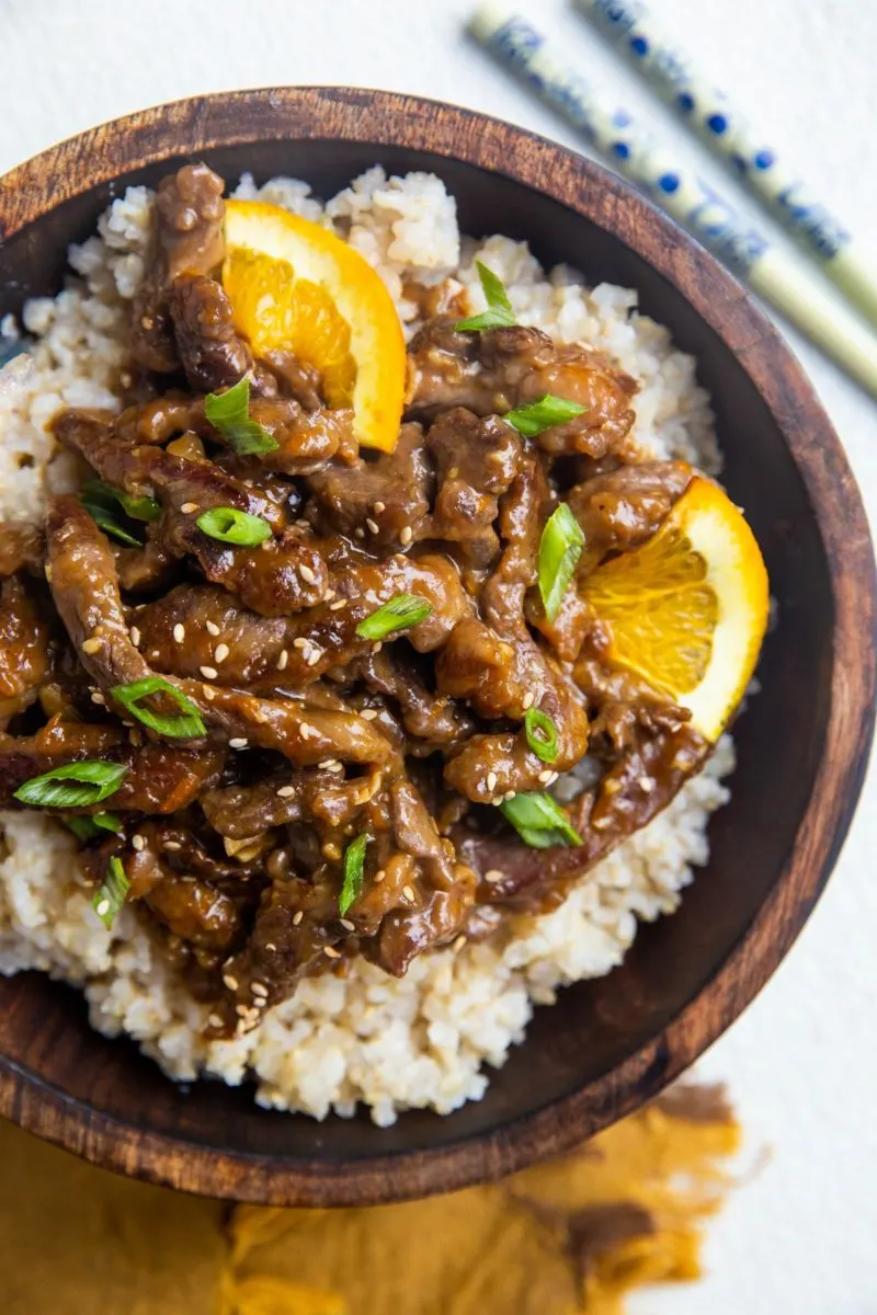 Wooden bowl of orange beef on top of brown rice with slices of orange. Chop sticks and a golden napkin to the side.