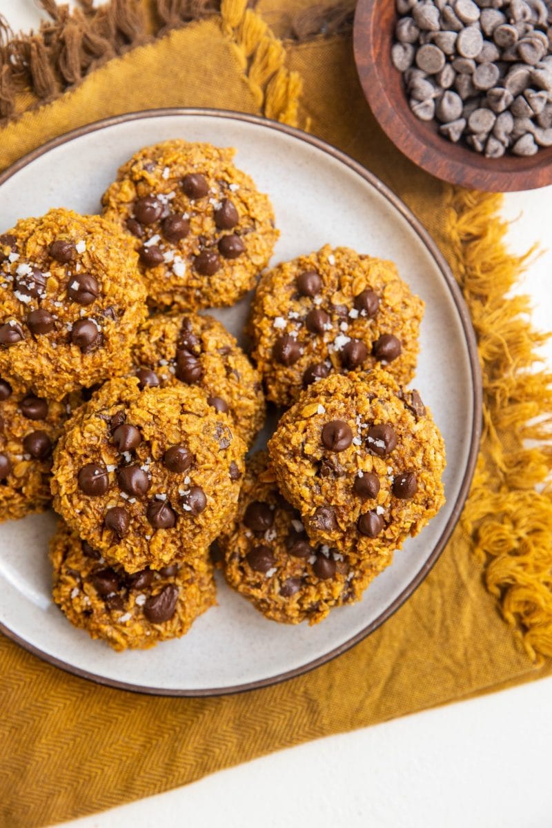 Plate of vegan oatmeal pumpkin cookies on top of a golden napkin with a bowl of chocolate chips to the side.