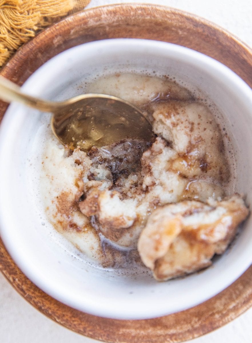 Close up top down image of cinnamon roll in a mug with a spoon digging in.