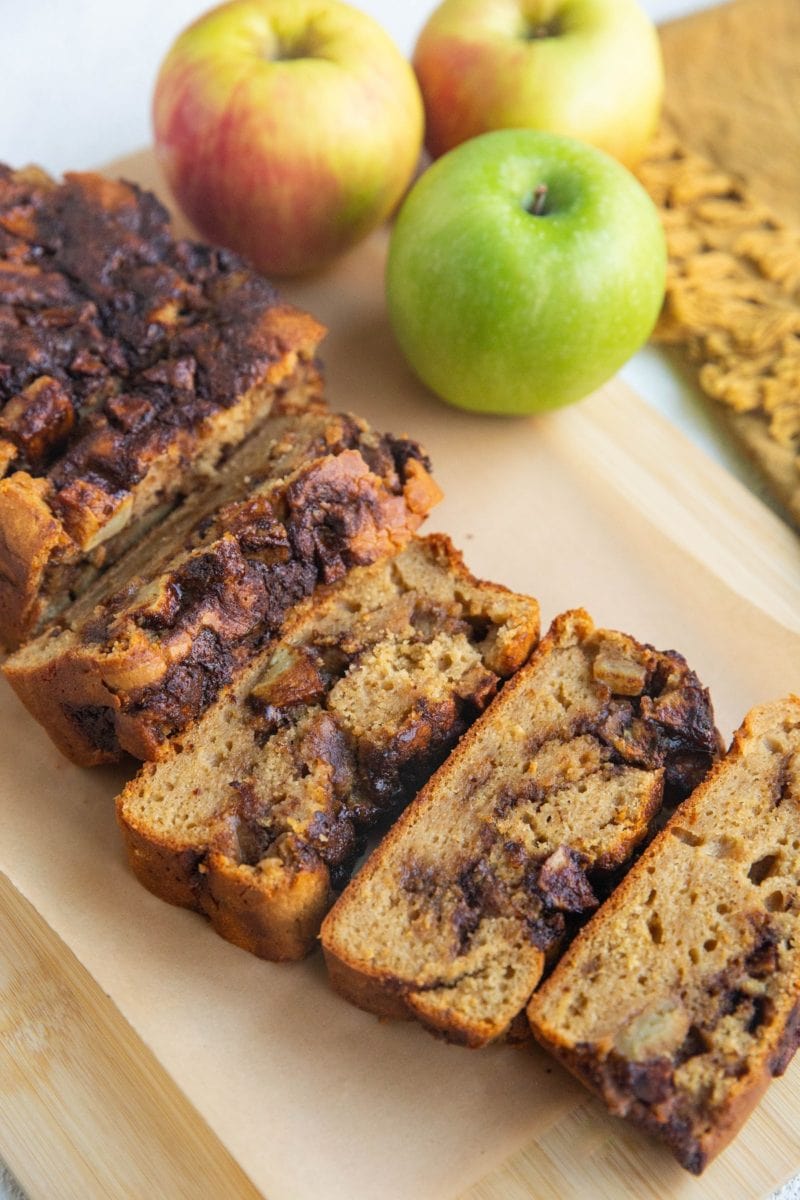 Loaf of apple cinnamon bread on a cutting board, cut into slices with fresh apples in the background.