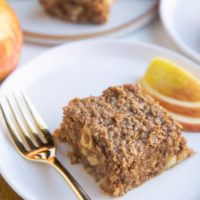 Slice of apple bar on a white plate with a gold fork and a plate of apple bars in the background.