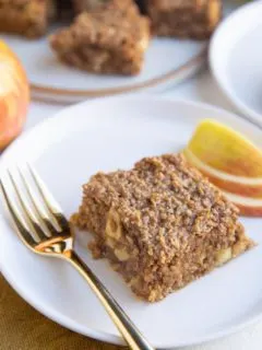 Slice of apple bar on a white plate with a gold fork and a plate of apple bars in the background.
