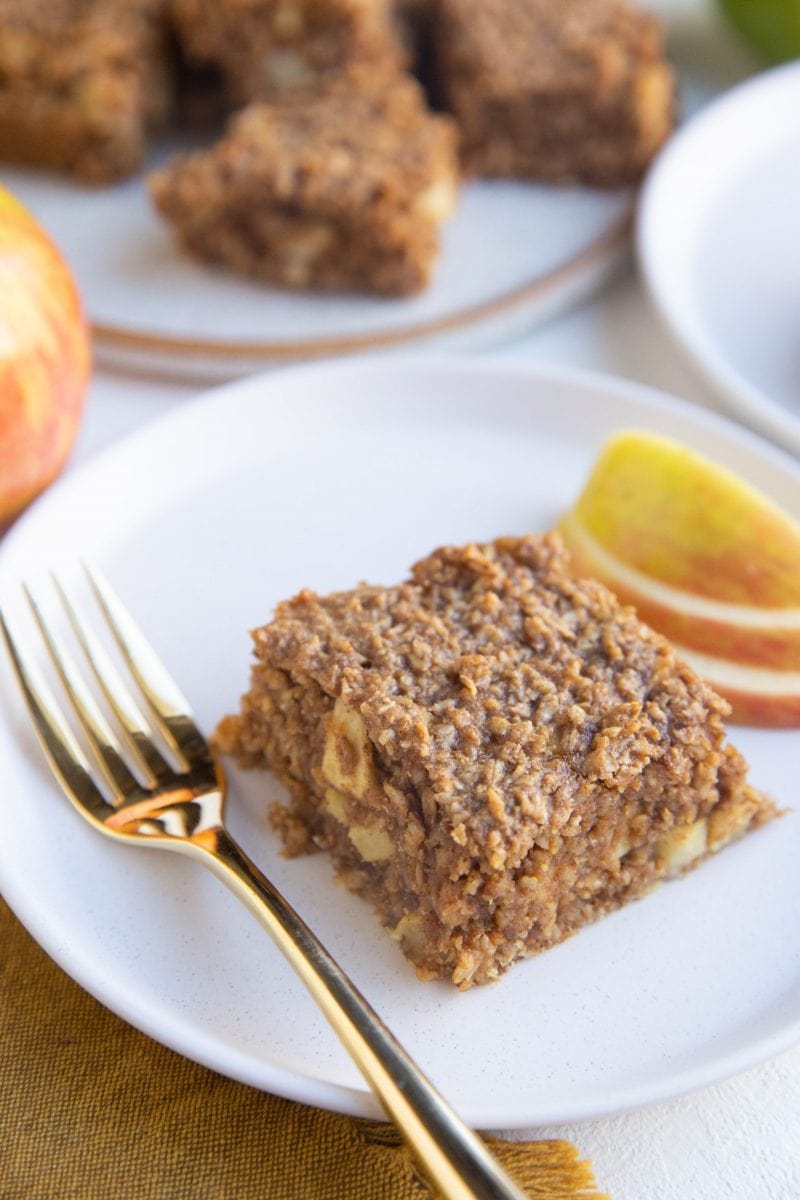 Slice of apple bar on a white plate with a gold fork and a plate of apple bars in the background.