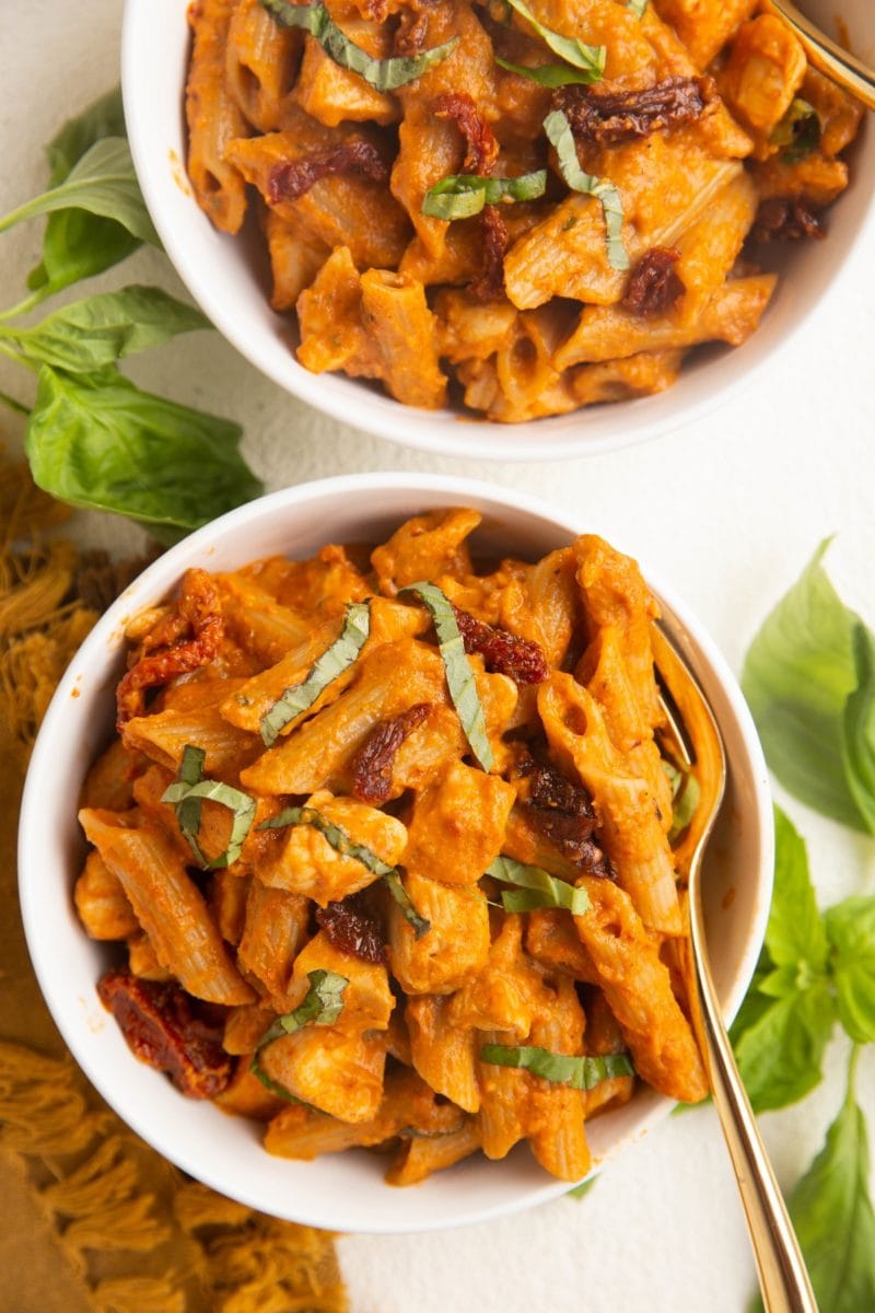 Two white bowls of pasta with a golden fork and fresh basil to the side.