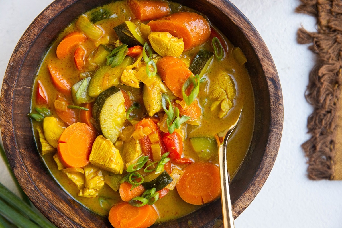 horizontal photo of wooden bowl of yellow curry soup with a gold spoon.