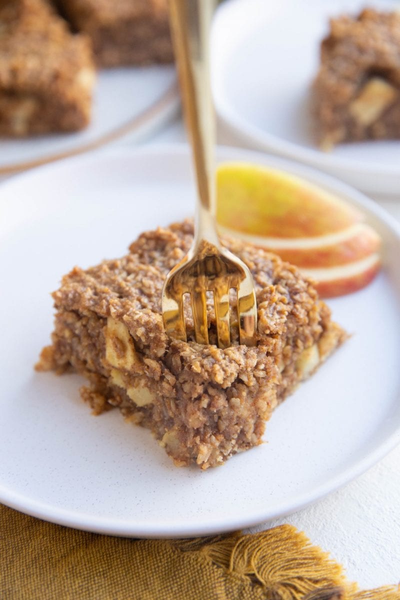 Slice of oatmeal bar on a white plate with a gold fork about to take a bite out.