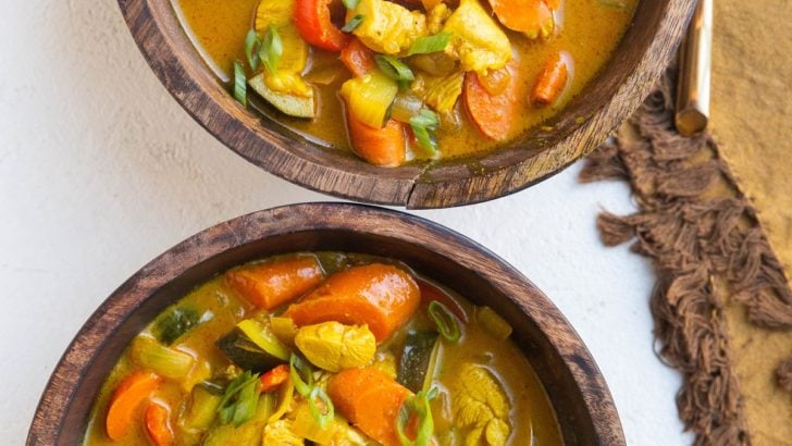 Two wooden bowls of yellow Thai curry soup with chicken and vegetables and gold spoons.