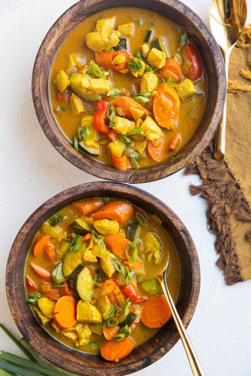 Two wooden bowls of yellow Thai curry soup with chicken and vegetables and gold spoons.