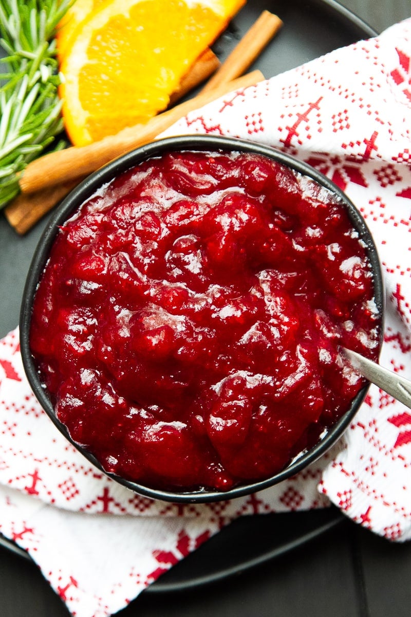 Black bowl of cranberry sauce with a napkin and sliced oranges and cinnamon sticks to the side.