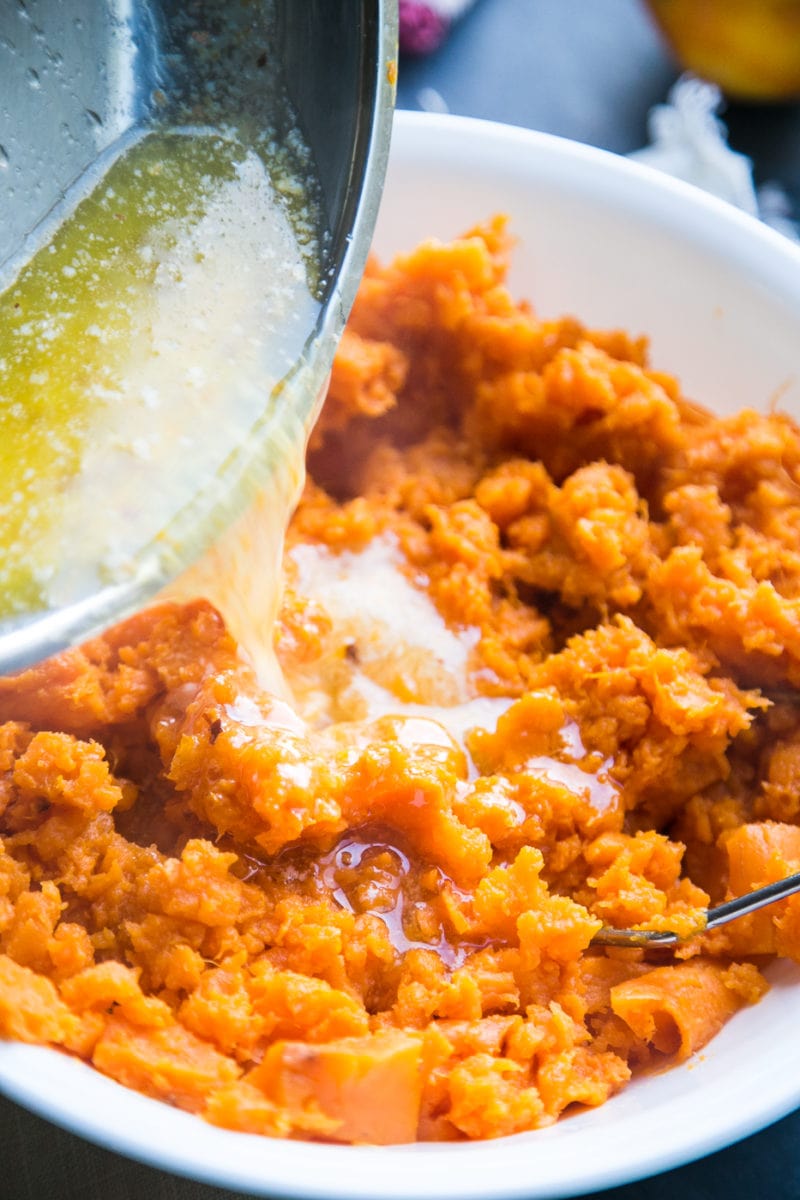 White bowl of mashed sweet potatoes with melted butter being poured into it.