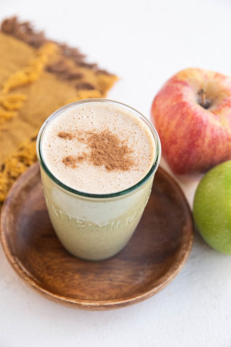 An apple pie smoothie in a glass with fresh apples to the side and a golden napkin.