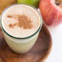 An apple pie almond butter smoothie in a glass with cinnamon on top and fresh apples in the background.