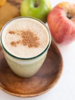An apple pie almond butter smoothie in a glass with cinnamon on top and fresh apples in the background.