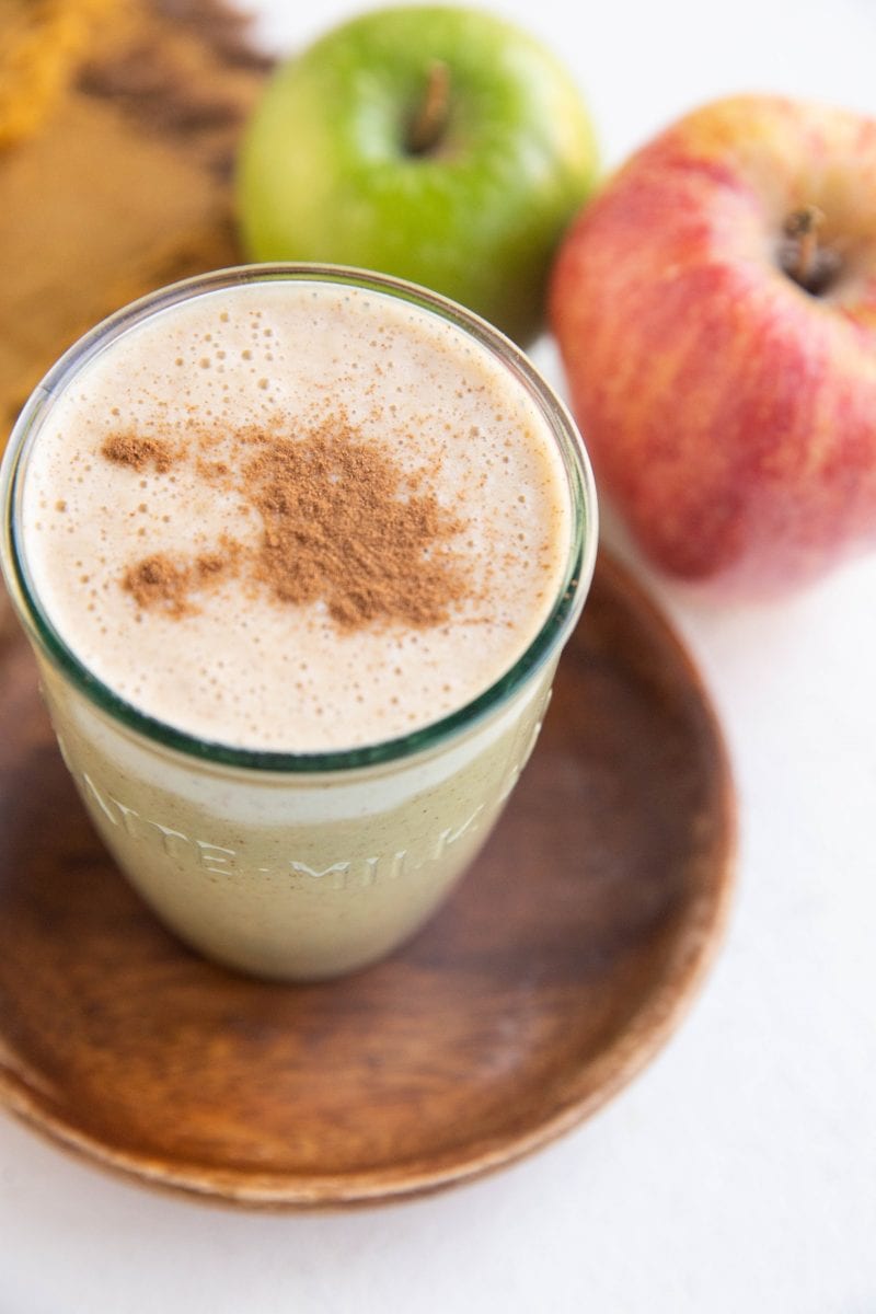 An apple pie almond butter smoothie in a glass with cinnamon on top and fresh apples in the background.