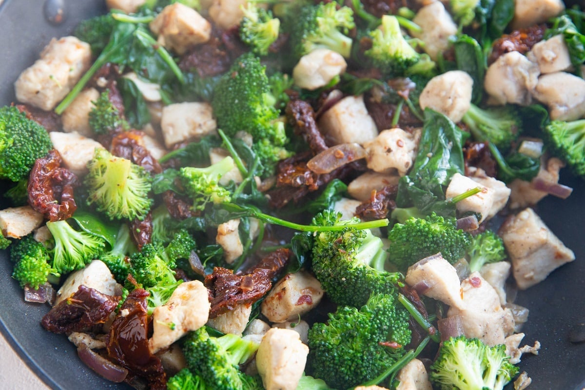 Onions, chicken, garlic, broccoli, sun-dried tomatoes and spinach cooking in a skillet.